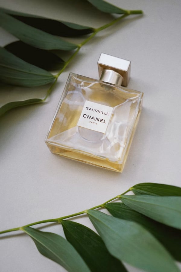 A bottle of Chanel Gabrielle perfume lying on a light surface, surrounded by green leaves.